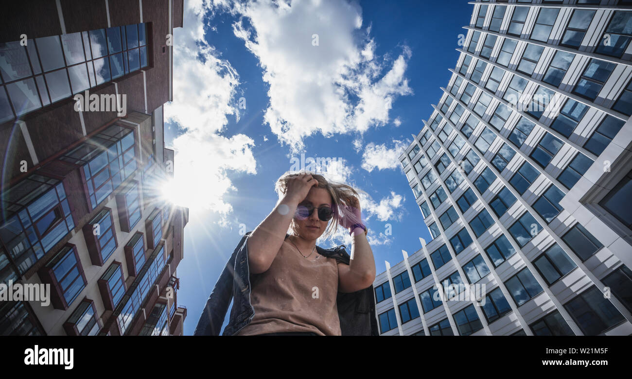 Portrait d'en bas contre le ciel bleu et les hautes maisons. Blonde à lunettes avec des cheveux ébouriffés. Le concept de la mode urbaine. Banque D'Images