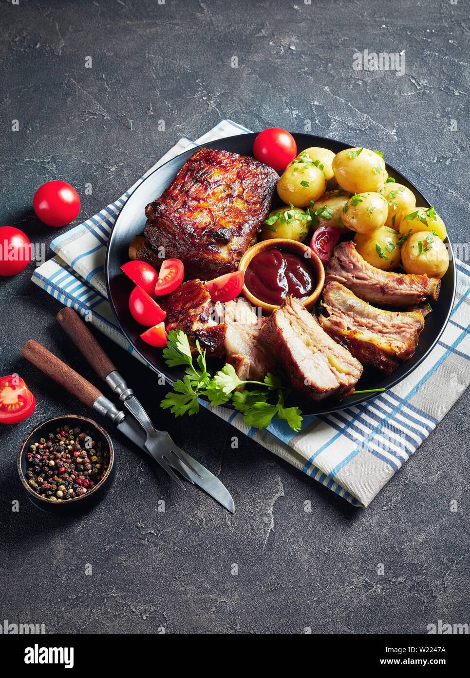 Côtes de porc grillées recouvertes de sauce barbecue servi avec des tomates et de l'eau chaude les pommes de terre servies sur un plateau noir sur une table, vue verticale Banque D'Images