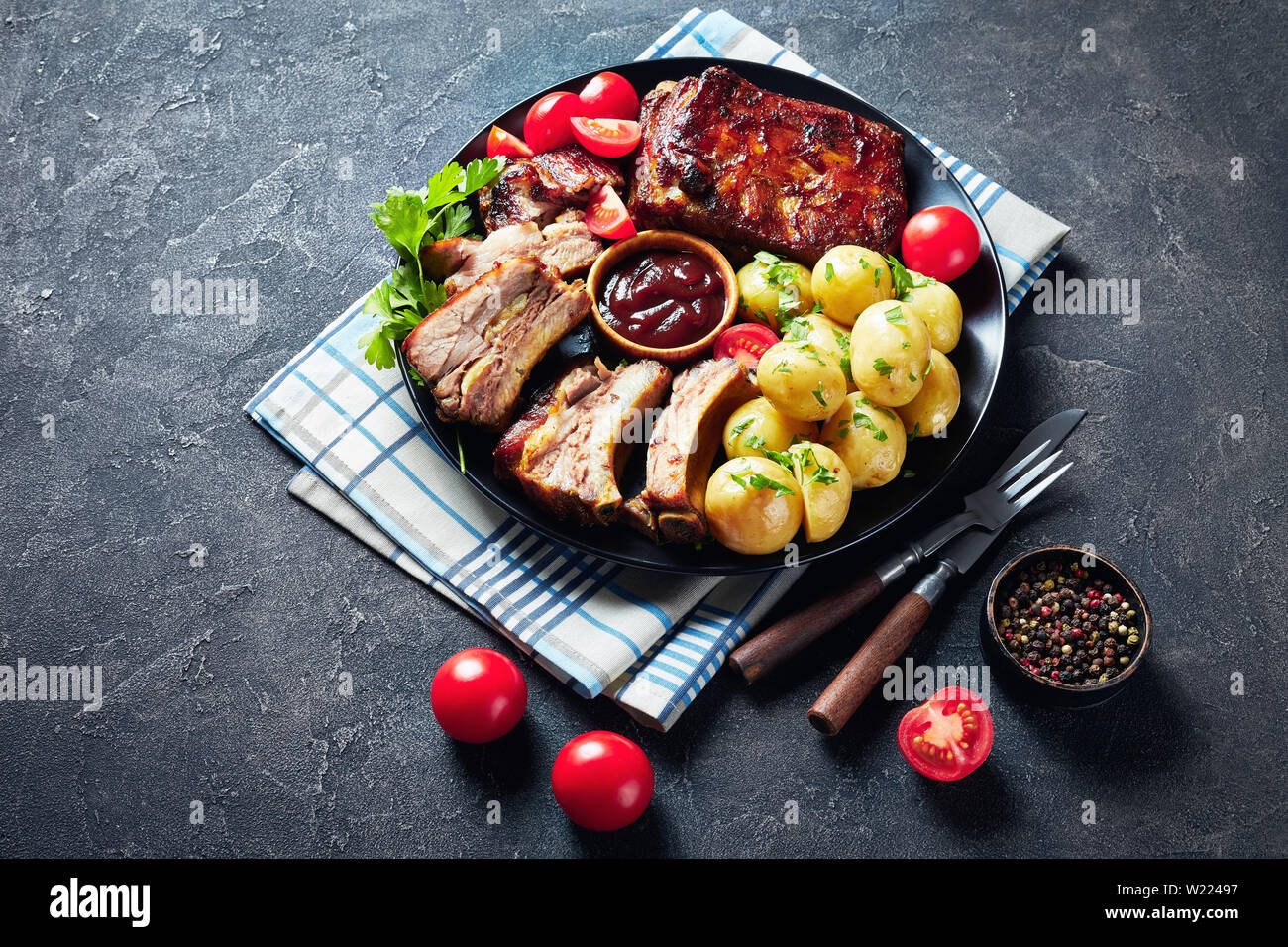 Côtes de porc grillé juteux délicieux garni de sauce barbecue servi avec des tomates et de l'eau chaude les pommes de terre servies sur un plateau noir sur une tabl Banque D'Images