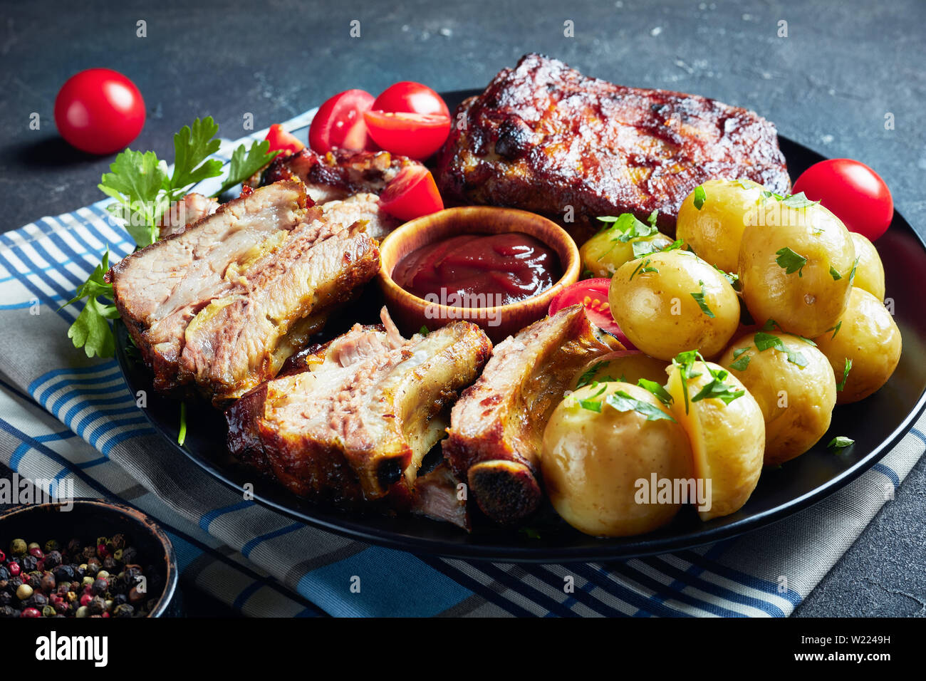 Côtes de porc grillées recouvertes de sauce barbecue servi avec des tomates et de l'eau chaude les pommes de terre servies sur un plateau noir sur une table, une vue d'abo Banque D'Images