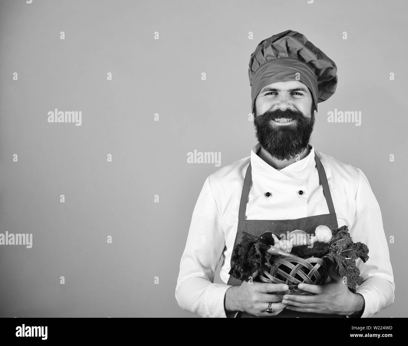 Cook avec visage souriant en bourgogne est titulaire d'uniforme des légumes dans un bol en osier. Chef détient la laitue, la tomate, le poivron et les champignons. Régime Végétarien concept. Homme à barbe sur fond rouge, copy space Banque D'Images