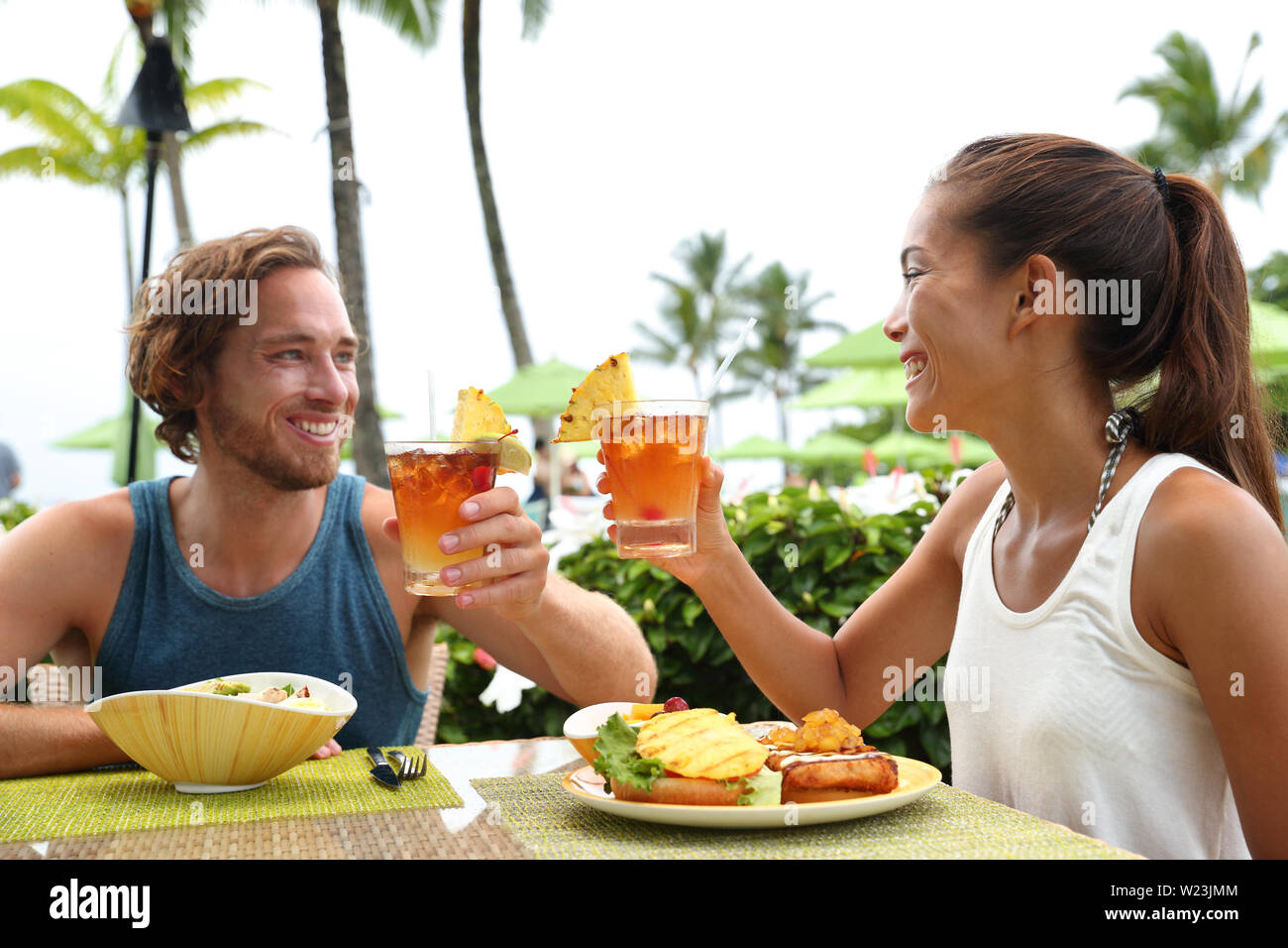 Heureux couple toasting multiraciale cheers avec boissons boissons hawaïenne, mai tai, Missouri l'expérience. Voyage Vacances d'été, les personnes bénéficiant de l'alimentation locale repas au restaurant terrasse extérieure de la station. Banque D'Images