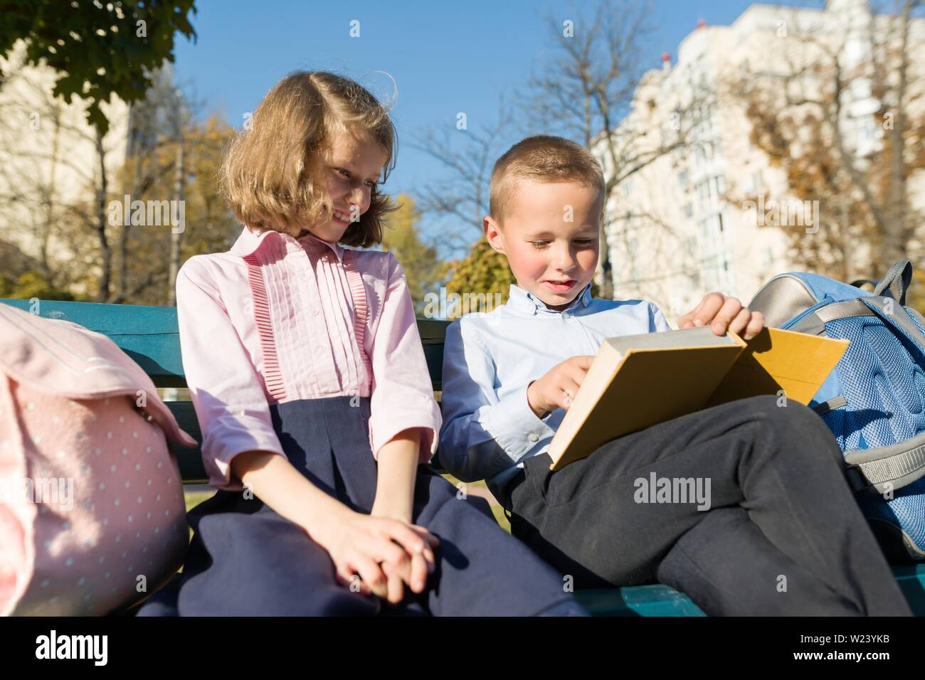 Petit garçon et fille écoliers lecture livre, assis sur un banc, des enfants avec des sacs à dos, journée d'automne ensoleillée Banque D'Images