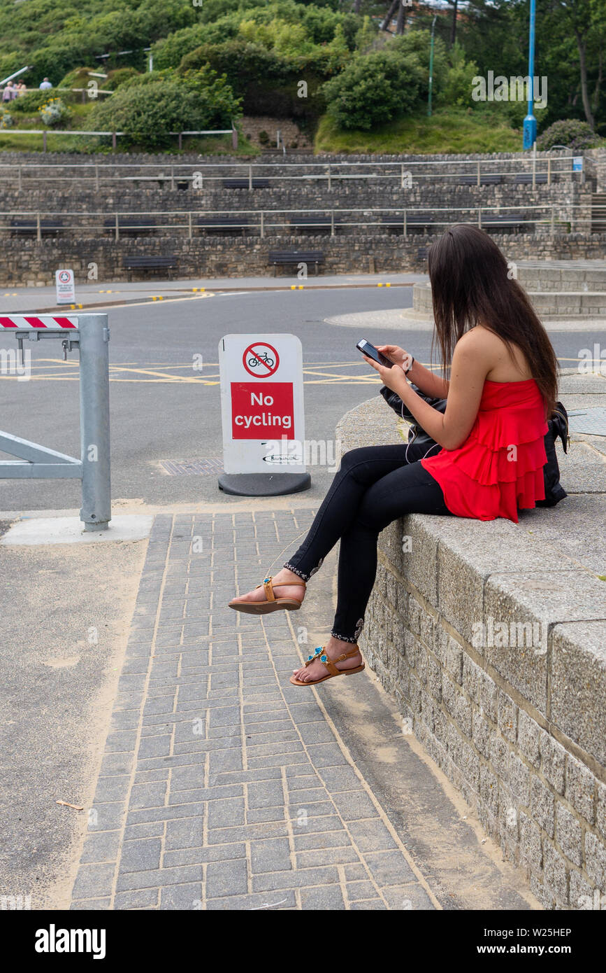 Jeune femme porter le rouge et le noir en utilisant un smartphone alors qu'il était assis à l'extérieur sur un mur. Banque D'Images