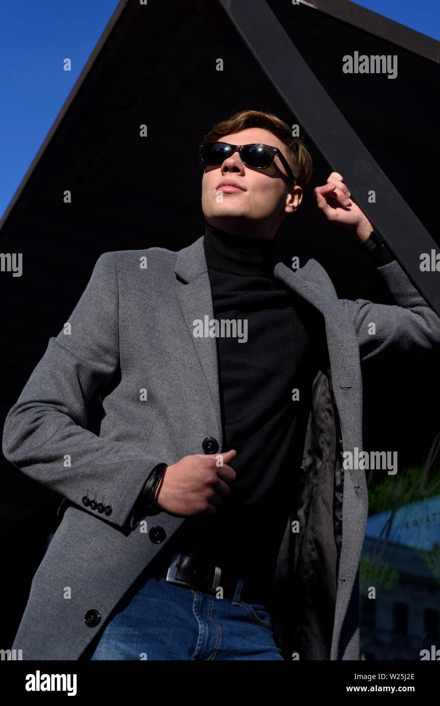 Homme élégant posant dans la lumière du soleil, vêtu de gris élégante veste, col polo noir et lunettes de soleil. Photo de mode vertical. Banque D'Images