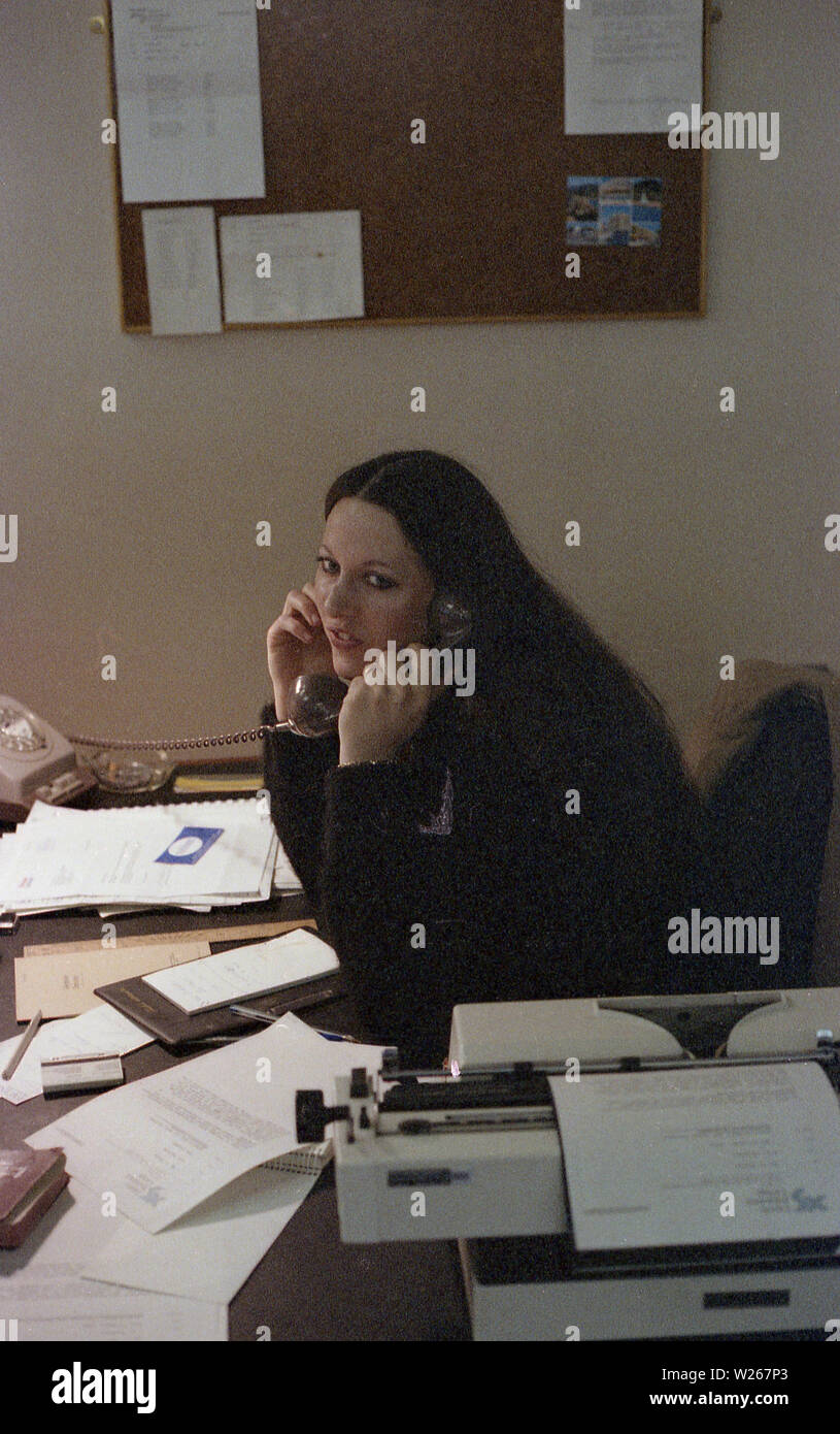 Années 1970, historiques, une jeune femme secrétaire ou employée de bureau, fin des années 20 dans l'âge, parlant au téléphone à son bureau, avec les formalités administratives et de machine à écrire. Banque D'Images