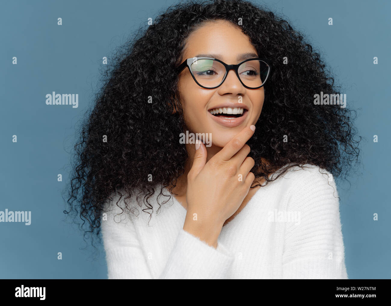 Très gros plan de la peau foncée agréable à la jeune femme est titulaire d'chin, regarde ailleurs, a les cheveux bouclés, large sourire, être en bonne humeur, isolé sur zone bleue Banque D'Images