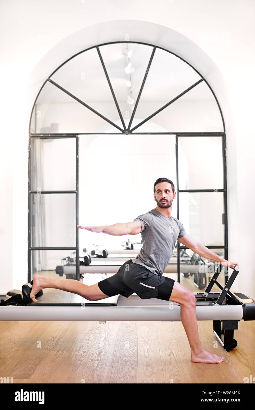 Un homme debout sur une jambe à l'aide d'un stretch pilates reformer lit dans une salle de sport à étirer ses fléchisseurs de hanche dans un concept de santé et de remise en forme Banque D'Images