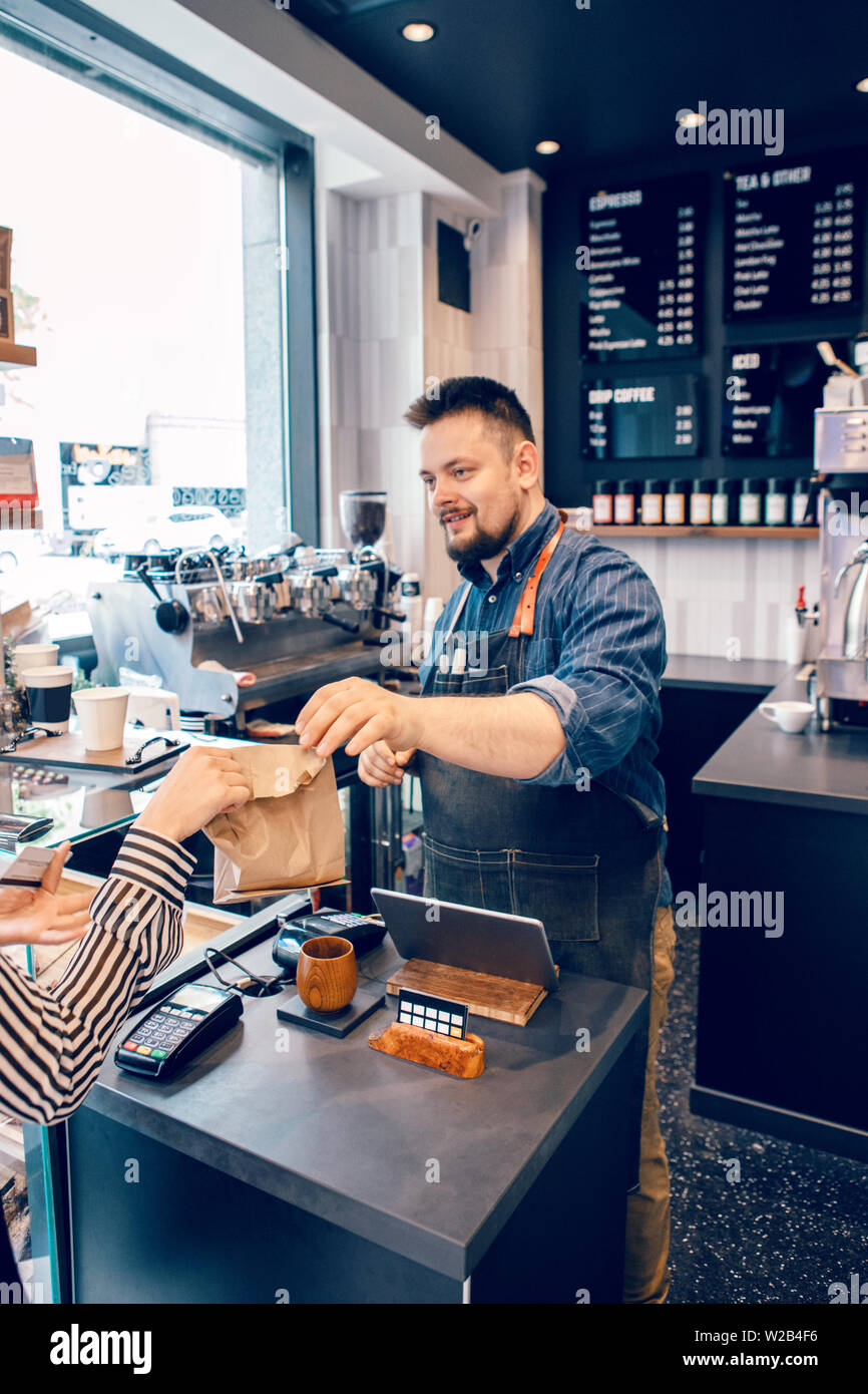 Smiling Caucasian man barbu en restaurant café barista caissier au travail. Vendeur client donnant son client dans l'achat de produits alimentaires biologiques packa marron papier Banque D'Images