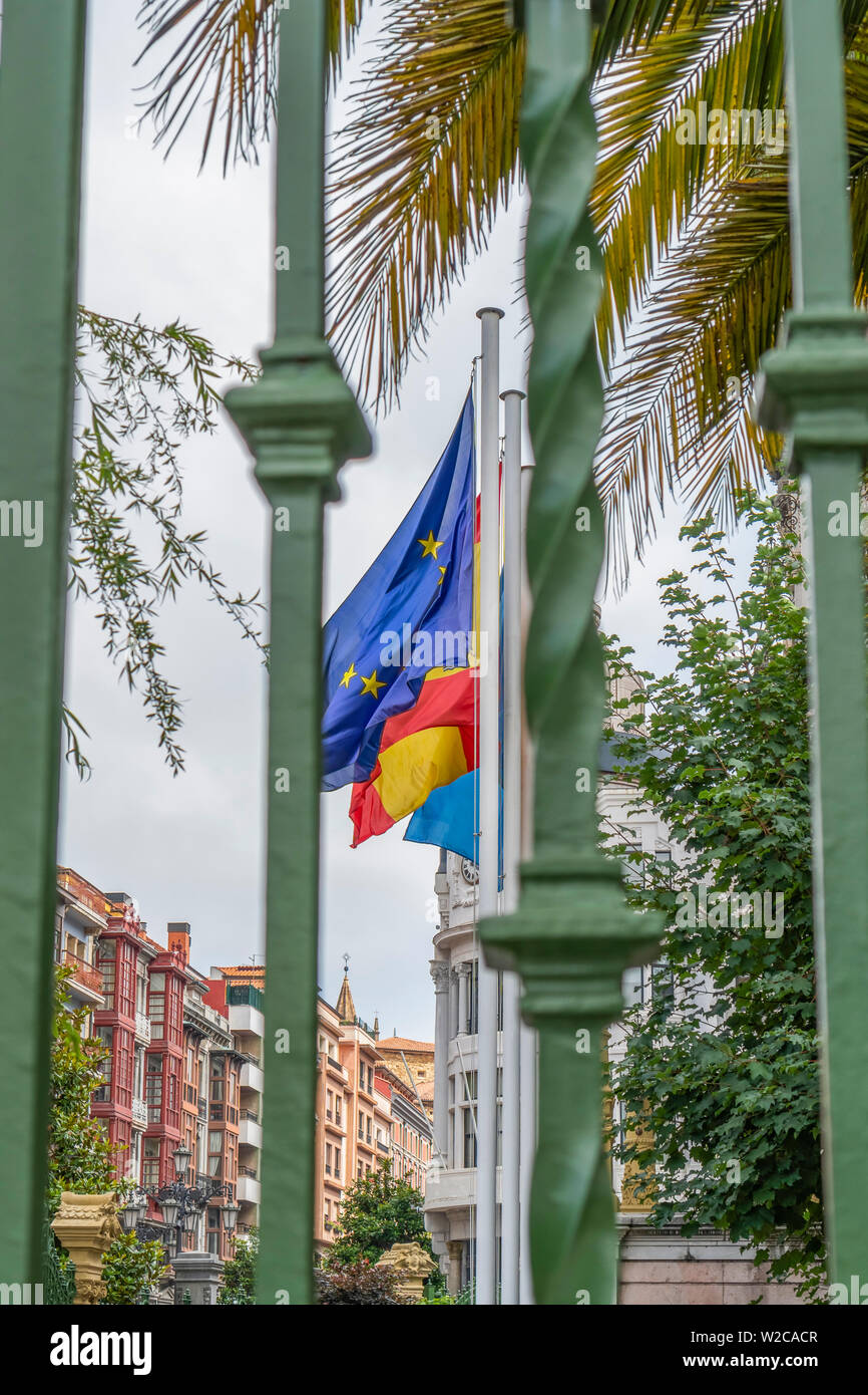 Drapeaux de l'Union européenne, l'Espagne, région des Asturies sur la façade d'un bâtiment ancien, derrière une grille de clôture verte. Contre les branches vertes de palm Banque D'Images