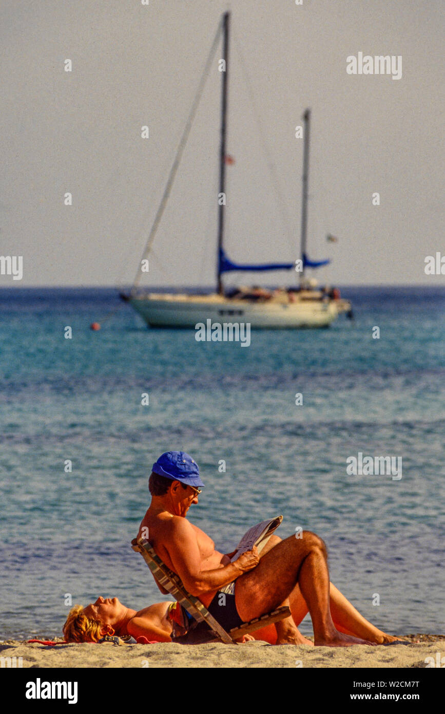 La plage de Pevero, Sardaigne, Italie. Un couple de soleil. L'homme lit un journal. Location de bateaux à l'ancre dans la mer derrière eux. Photo : © Simon Grosset. Archive : image numérisé à partir d'un original de la transparence. Banque D'Images