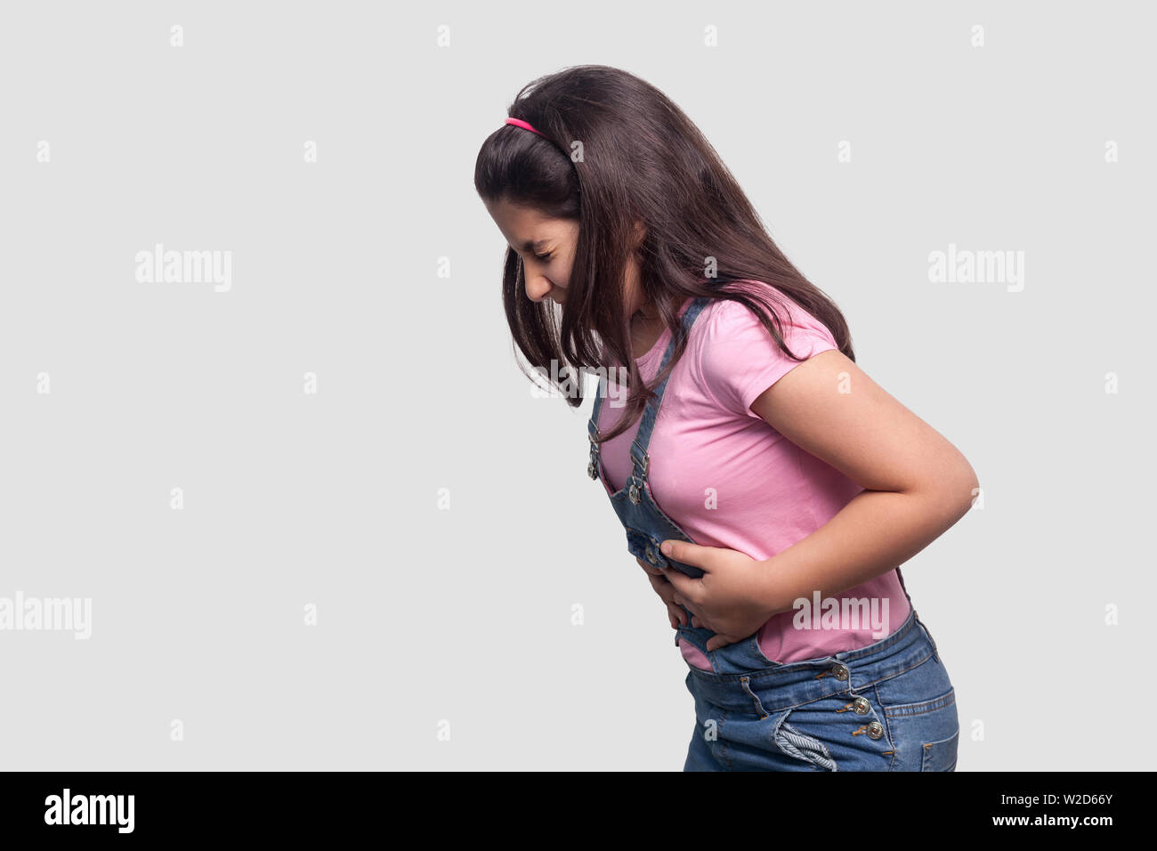 La douleur d'estomac. Portrait de profil Vue de côté d'un brunette jeune fille en rose t-shirt et salopette bleue debout et tenant son ventre malade douloureux. en Banque D'Images