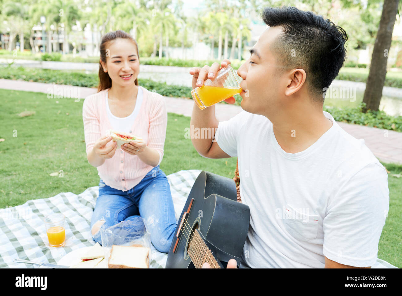 Young Asian man with guitar de boire du jus d'orange avec femme assise près de lui et maintenant ils sont en sandwich pique-nique dans le parc Banque D'Images