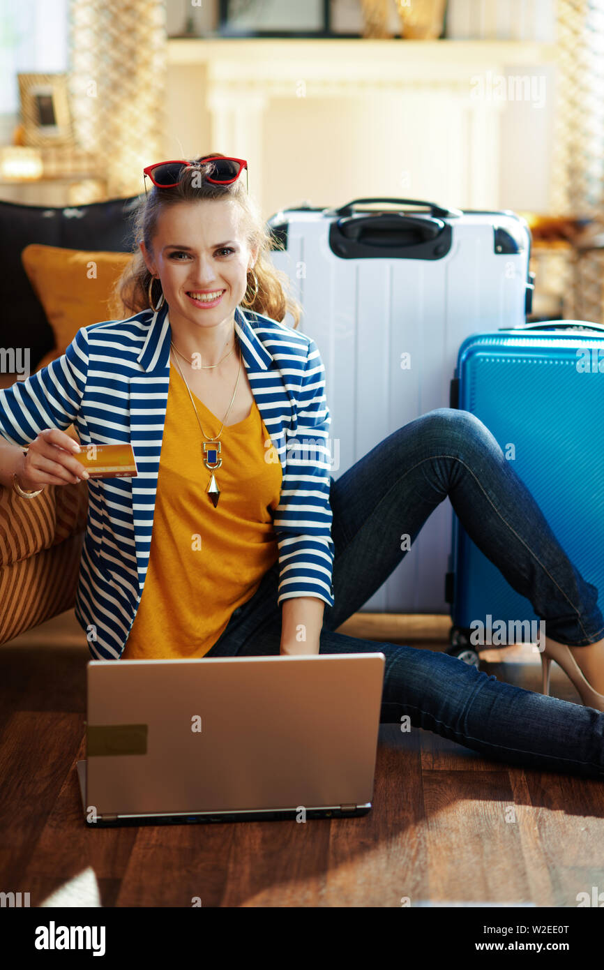 Happy voyageur élégant femme en blouson rayé avec carte de crédit l'achat des billets d'avion en ligne sur un ordinateur portable tout en étant assis près de la table et l'sacs trolley Banque D'Images