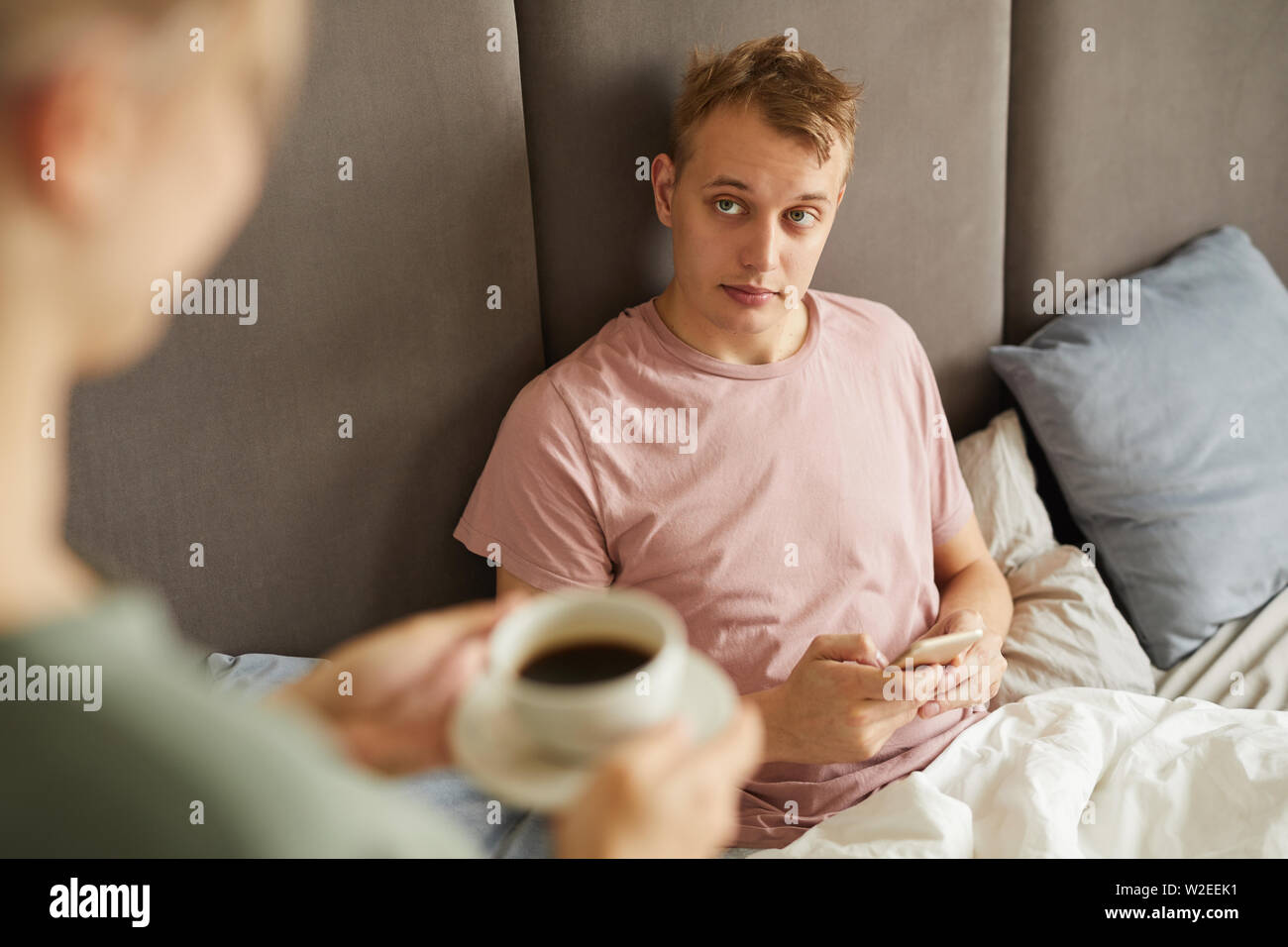 Jeune homme fatigué ou malade avec le smartphone à son épouse à l'emmenant tasse de café au lit Banque D'Images
