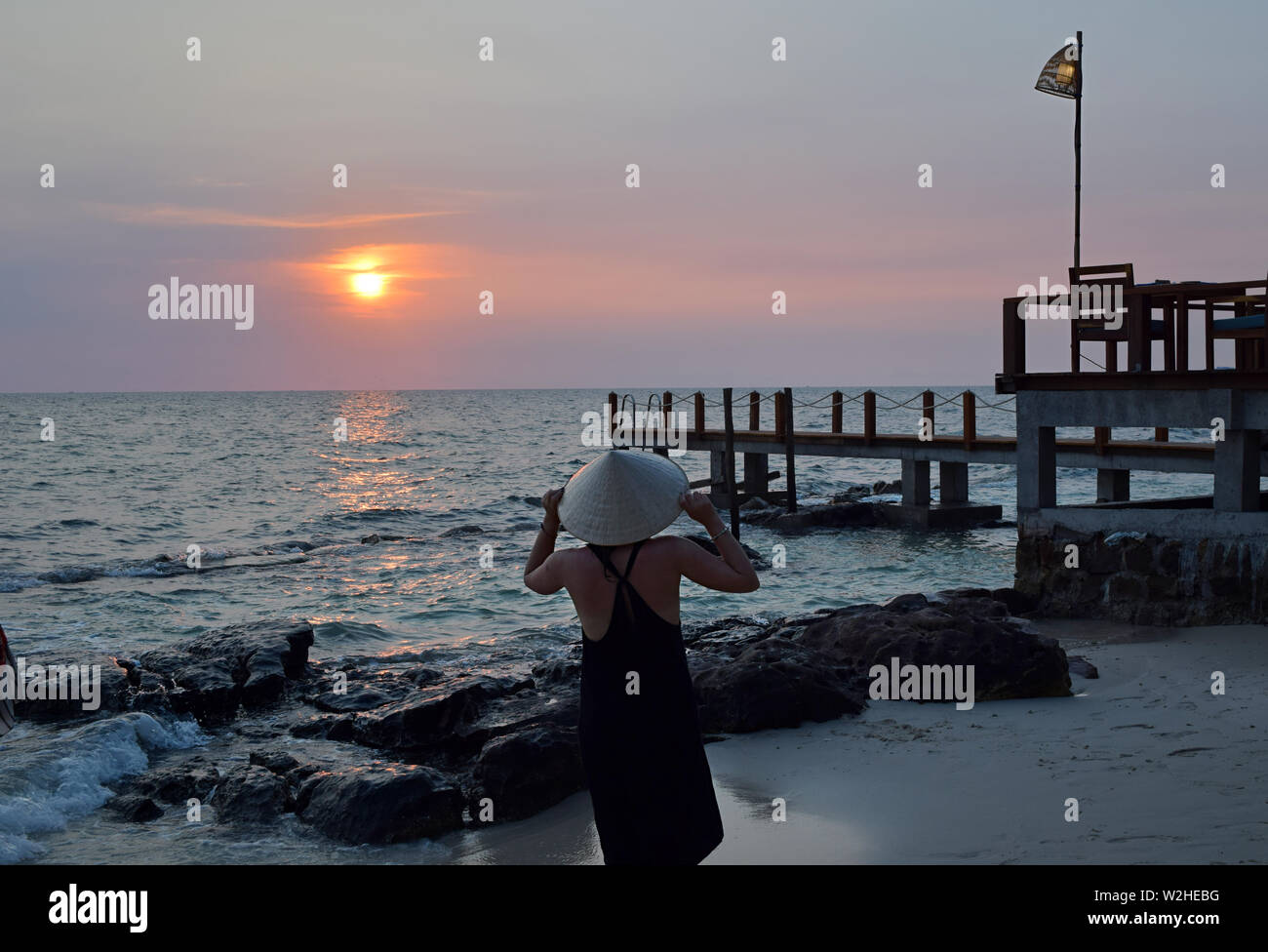 Jeune femme séduisante au bord de la mer dans le port d'un chapeau typique du Vietnam à regarder le coucher du soleil. Jeune femme regardant le coucher du soleil sur la mer de Chine du Sud dans la jetée. Banque D'Images