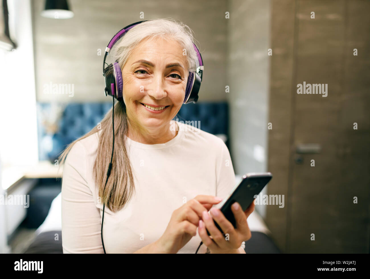 Hauts femme aux cheveux blancs dans la recherche d'écouteurs chansons préférées sur le smartphone et souriant à l'appareil photo Banque D'Images