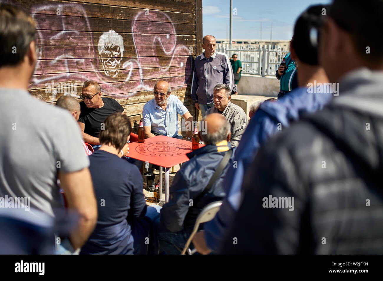 Bari, Italie - 05 mai 2019 : groupe d'hommes âgés de l'italien est relaxant avec de la bière par la table rouge sur la ville ensoleillée street. Ils portent des vêtements décontractés Banque D'Images