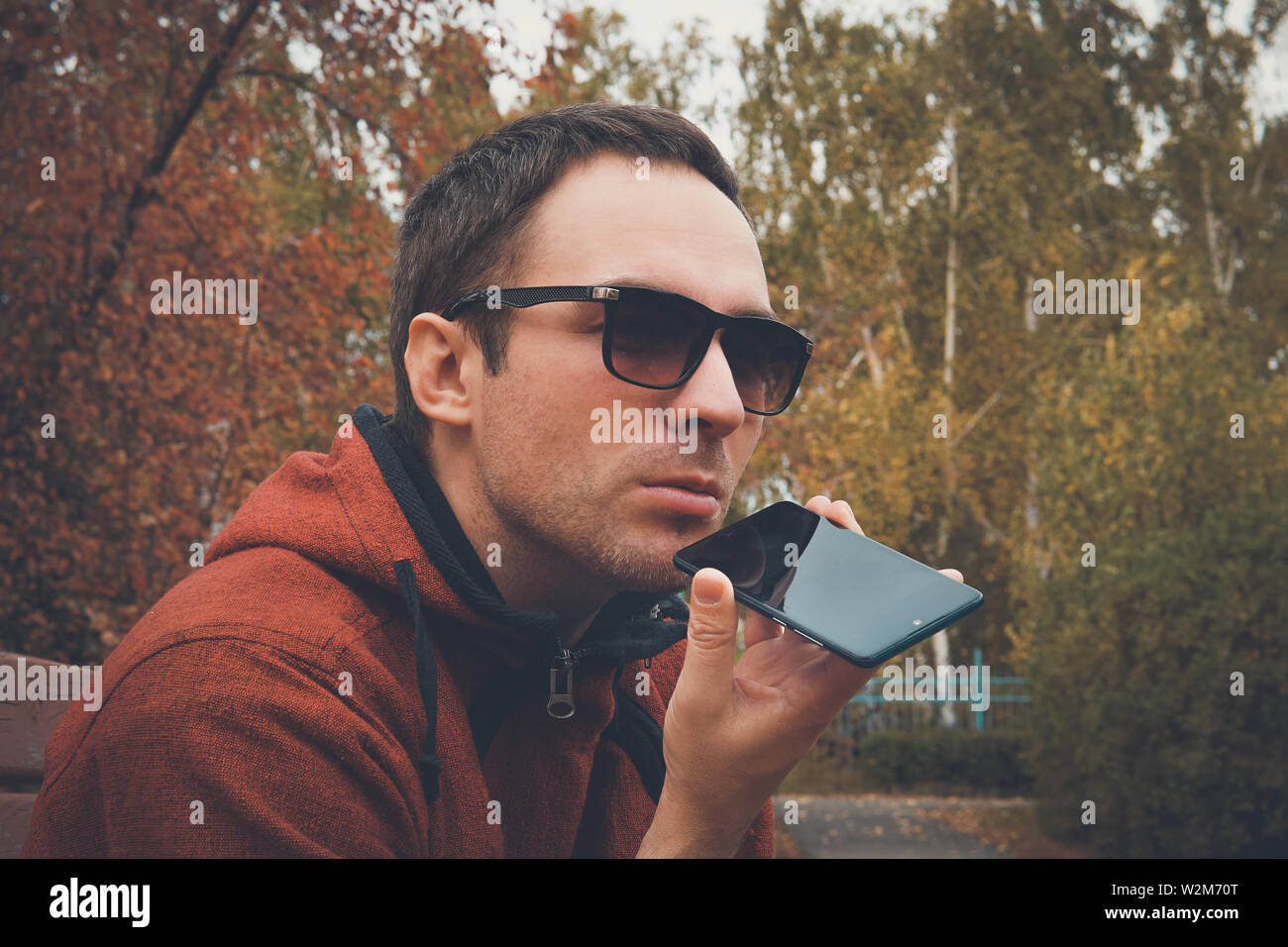 Beau jeune homme debout dans le parc en été, à l'aide d'un téléphone cellulaire à l'écoute de message vocal enregistré ou audio. guy parle dans le microphone à Banque D'Images