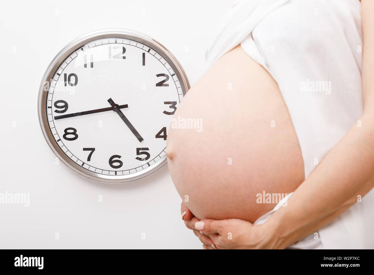 Pregnant woman holding her belly au cours des dernières semaines de la grossesse l'article par horloge de mur blanc, se concentrer sur l'horloge. Temps d'attente et concept Banque D'Images