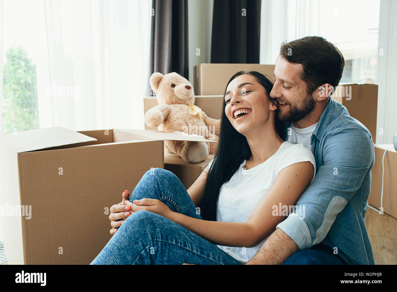 Heureux Couple hugging and sitting on floor dans leur nouvelle maison avec de nombreux cartons on background Banque D'Images