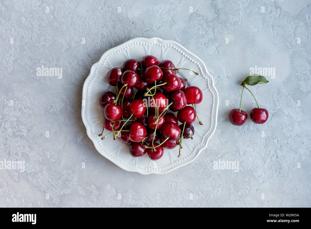Mise à plat avec plaque de cerises sur fond de béton gris. Concept de l'alimentation d'été. Vue de dessus, de l'espace pour texte et logo Banque D'Images Mise à plat avec plaque de cerises sur fond de béton gris. Concept de l'alimentation d'été. Vue de dessus, de l'espace pour texte et logo Banque D'Images