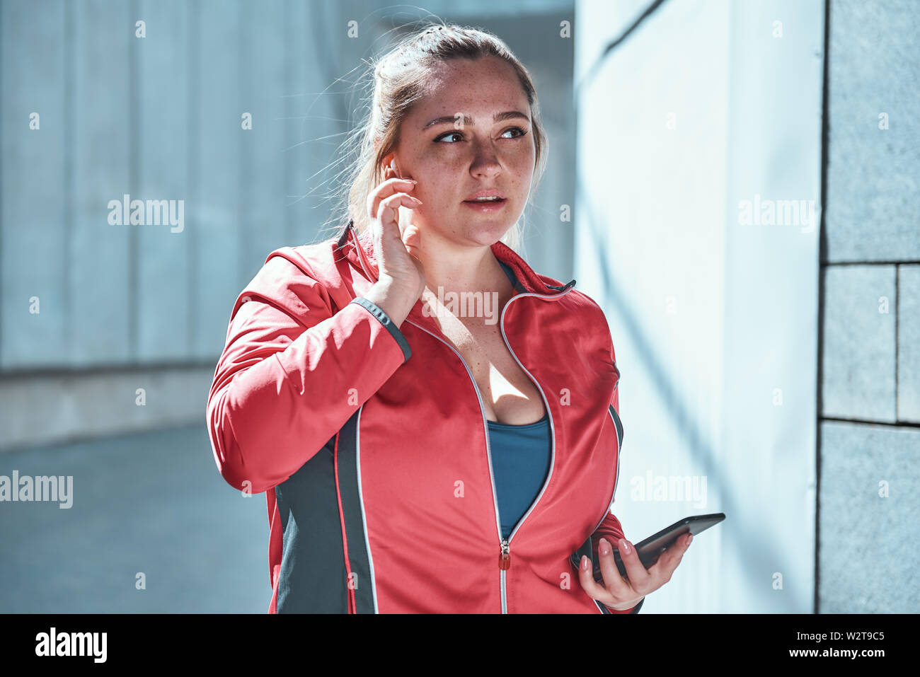 Taille plus femme attrayante dans les écouteurs holding smart phone et l'écoute de la musique tout en se tenant contre un mur extérieur gris Banque D'Images
