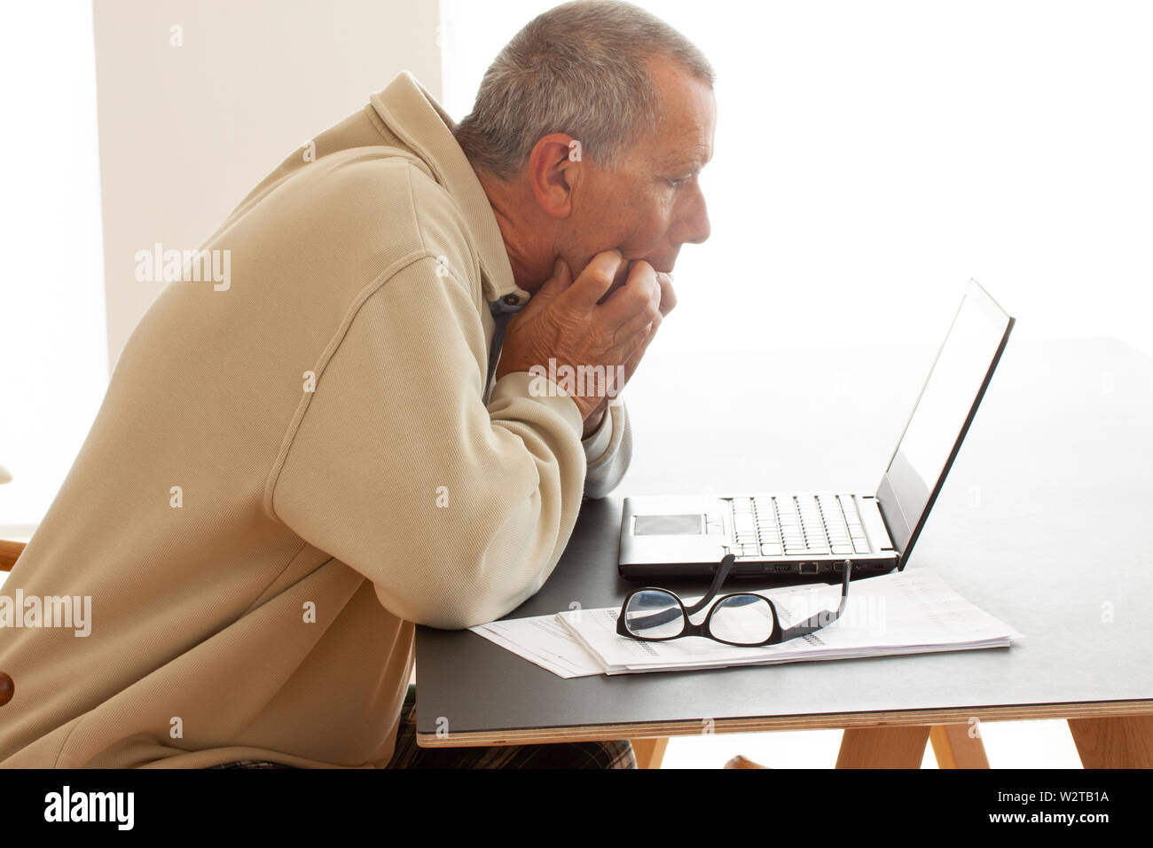 Habillé décontracté et anxieux man biting ses doigts comme il regarde un écran d'ordinateur. Une paire de lunettes est posé sur une pile de documents à côté de l'ordinateur portable com Banque D'Images