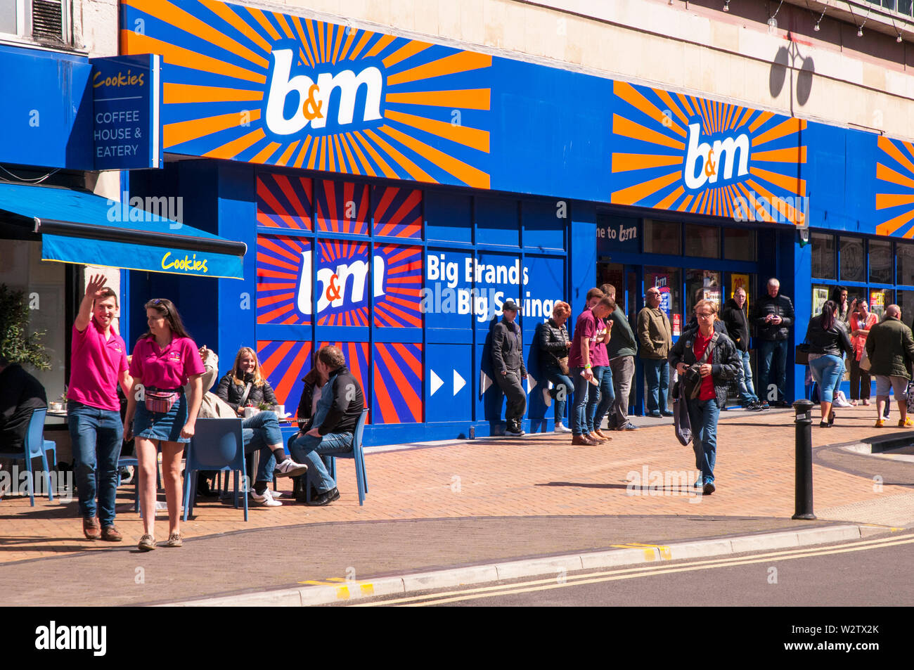 Hommes debout à l'extérieur B&M Bargain store possible en attente de femmes/copines à terminer shopping People sitting Café à l'extérieur et l'homme en agitant à l'appareil photo Banque D'Images