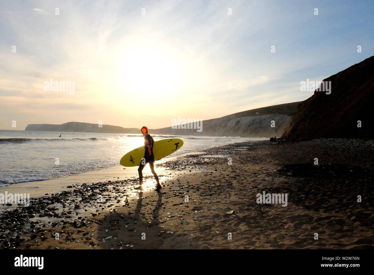Un jeune homme portant sa planche de surf sous le bras tête dans la mer à pied avec le soleil couchant et Tennyson en bas des falaises de craie dans l'arrière-plan Banque D'Images