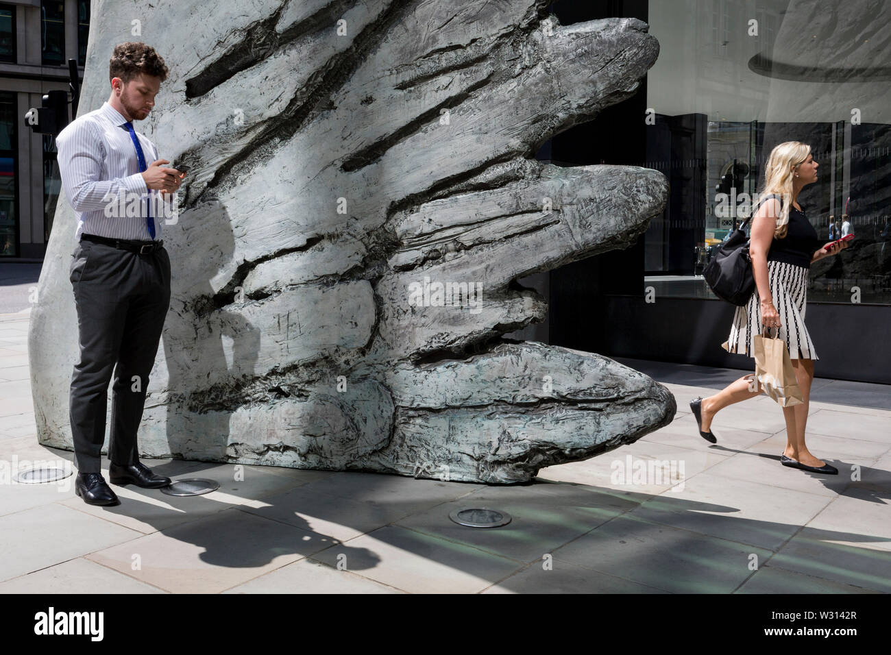 Un homme d'affaires de l'industrie financière vérifie les messages comme une dame marche l'autre côté de la sculpture intitulée City Wing sur Threadneedle Street dans la ville de Londres, le quartier financier de la capitale (aka le Square Mile), le 11 juillet 2019, à Londres, en Angleterre. L'aile est de la ville par l'artiste Christopher Le Brun. Les dix mètres de haut est la sculpture en bronze par le président de la Royal Academy of Arts, Christopher Le Brun, commandé par Hammerson en 2009. Elle est appelée 'La Ville' de l'aile et a été jeté par Morris Singer, fondateurs de l'art réputé pour être la plus ancienne fonderie d'art dans le monde. Banque D'Images
