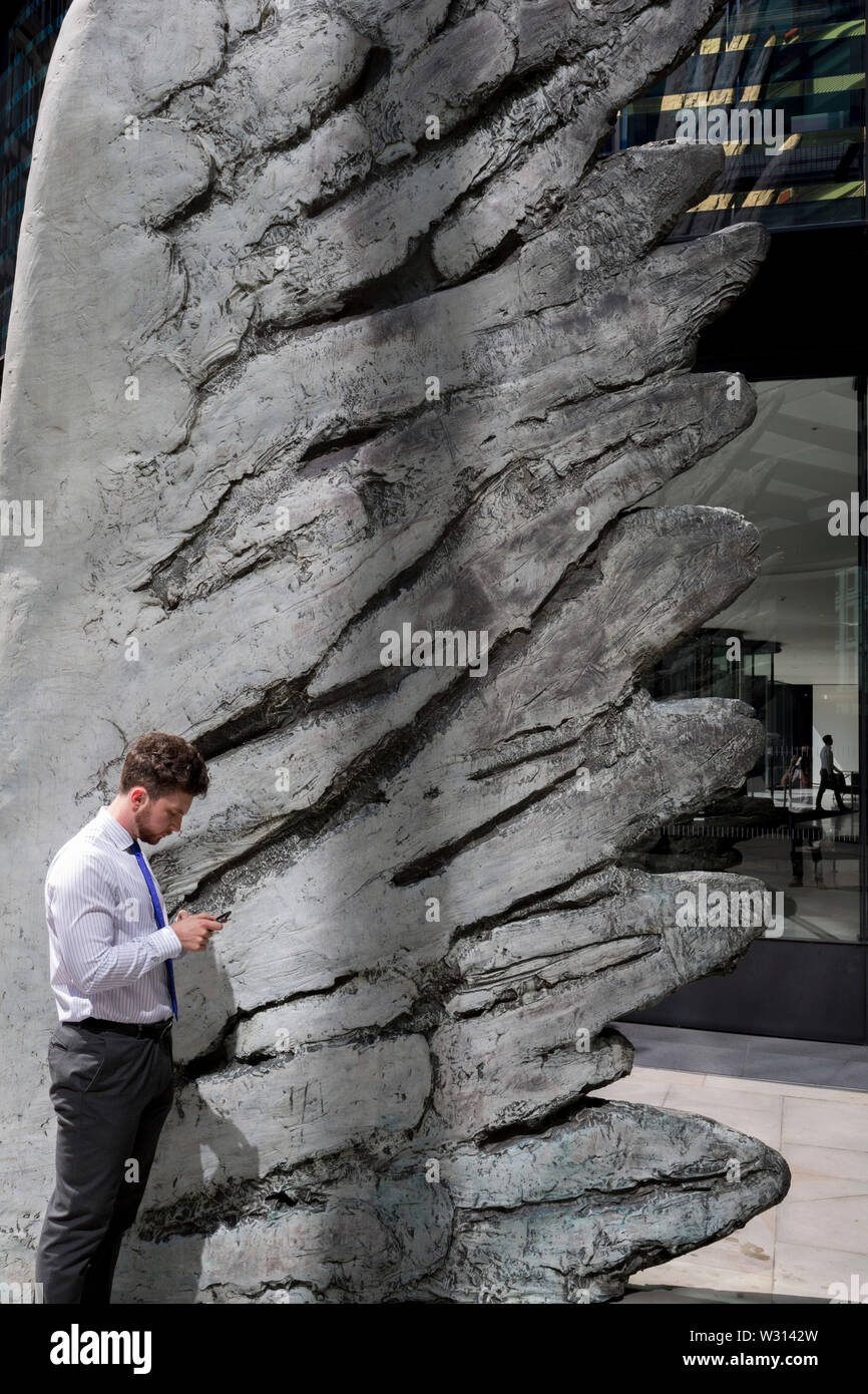 Un buusinessman l'industrie financière vérifie les messages sous la sculpture intitulée City Wing sur Threadneedle Street dans la ville de Londres, le quartier financier de la capitale (aka le Square Mile), le 11 juillet 2019, à Londres, en Angleterre. L'aile est de la ville par l'artiste Christopher Le Brun. Les dix mètres de haut est la sculpture en bronze par le président de la Royal Academy of Arts, Christopher Le Brun, commandé par Hammerson en 2009. Elle est appelée 'La Ville' de l'aile et a été jeté par Morris Singer, fondateurs de l'art réputé pour être la plus ancienne fonderie d'art dans le monde. Banque D'Images