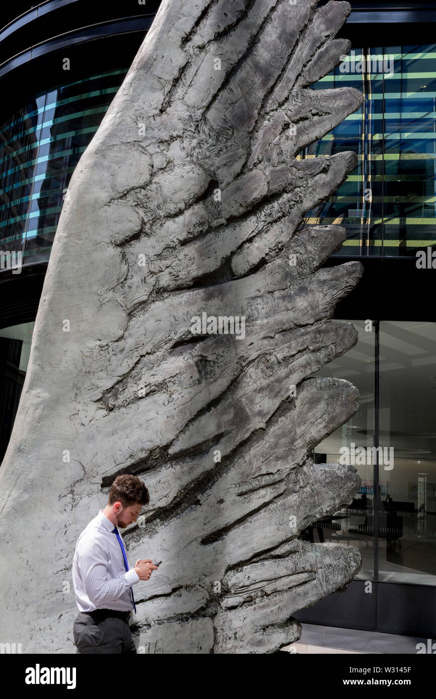 Un buusinessman l'industrie financière vérifie les messages sous la sculpture intitulée City Wing sur Threadneedle Street dans la ville de Londres, le quartier financier de la capitale (aka le Square Mile), le 11 juillet 2019, à Londres, en Angleterre. L'aile est de la ville par l'artiste Christopher Le Brun. Les dix mètres de haut est la sculpture en bronze par le président de la Royal Academy of Arts, Christopher Le Brun, commandé par Hammerson en 2009. Elle est appelée 'La Ville' de l'aile et a été jeté par Morris Singer, fondateurs de l'art réputé pour être la plus ancienne fonderie d'art dans le monde. Banque D'Images