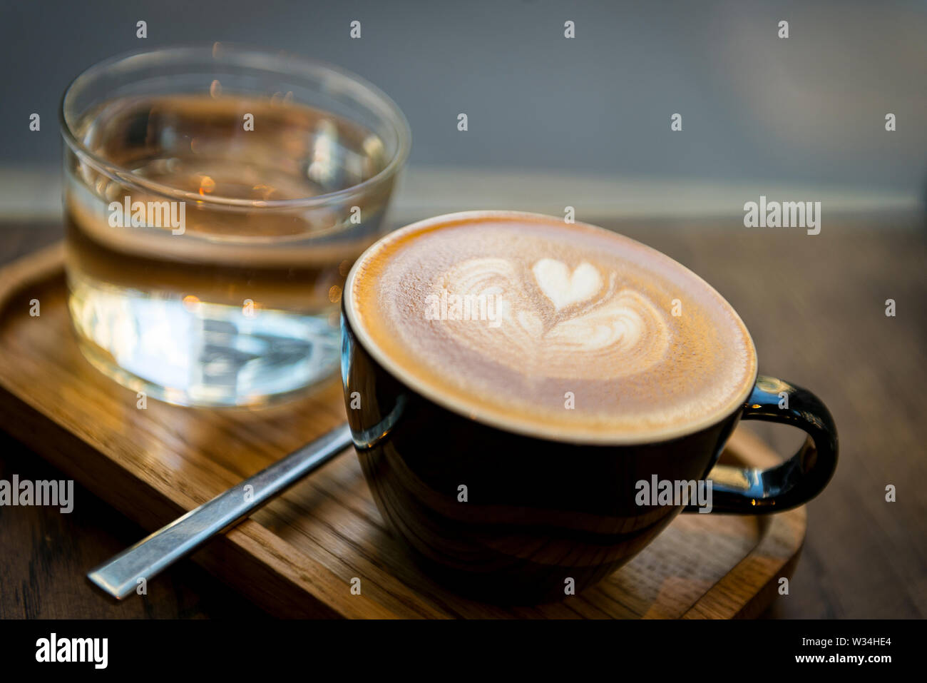Latte art dans une tasse noire sur un plateau en bois avec un verre d'eau et une cuillère ; café confortable gros plan d'une boisson expresso de spécialité pour une pause café du matin. Banque D'Images