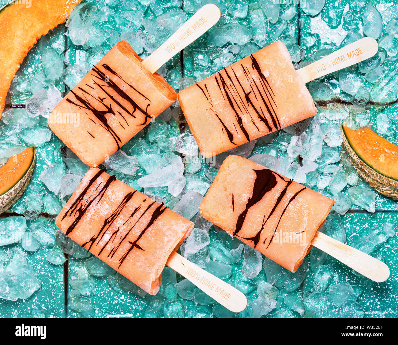 Vue de dessus des sucettes glacées faites de melon cantaloup avec du chocolat et des cubes de glace sur fond turquoise rustique. Banque D'Images