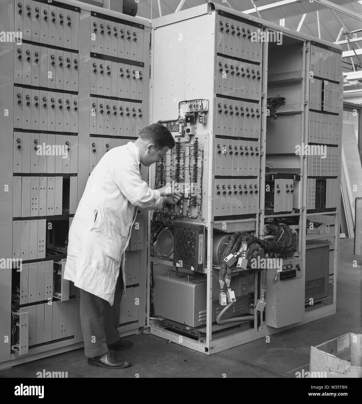 1960s, historique, un technicien masculin en blouse blanche travaillant dans l'industrie aéronautique sur l'équipement électrique, un tableau de distribution ou un tableau de bord, qui sépare l'alimentation secteur entrante en divers sous-circuits. Ces panneaux sont utilisés pour de grands volumes d'équipements électriques et sont vus dans les usines et les locaux industriels. Banque D'Images