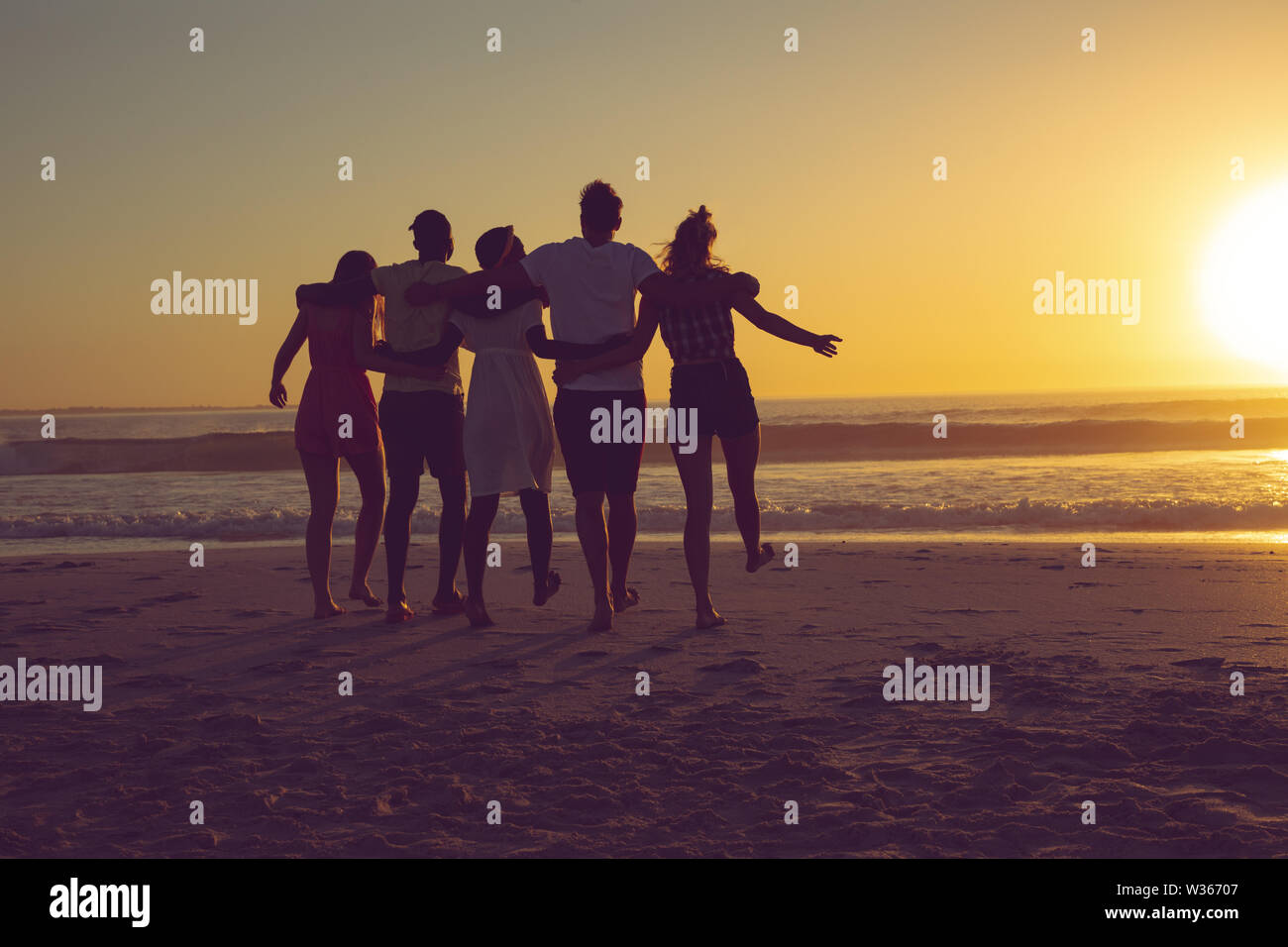 Amis avec bras autour de marcher vers la plage pendant le coucher du soleil Banque D'Images