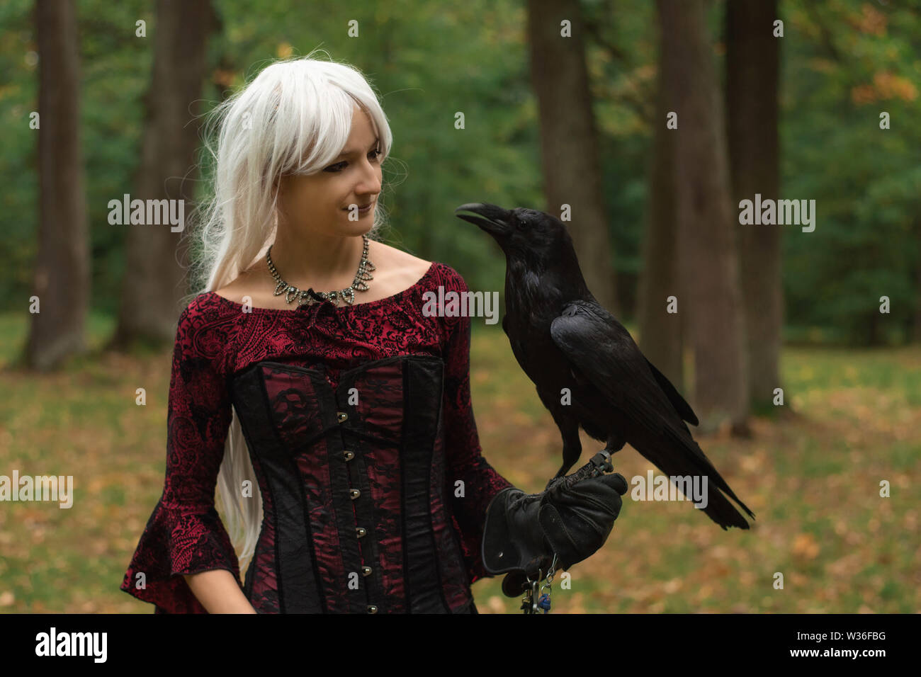 Sorcière femme en robe rouge et avec raven dans ses mains dans la forêt enneigée. Elle regarde Raven. Autour de neige et les flocons tombent sur l'ourlet de sa robe. Banque D'Images