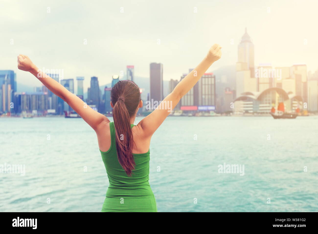 Happy woman cheering succès par Hong Kong city skyline with arms raised up tendus. Gagnant de célébrer le coucher du soleil, la promenade de Tsim Sha Tsui et de l'Avenue des Stars à Victoria Harbour, Kowloon. Banque D'Images