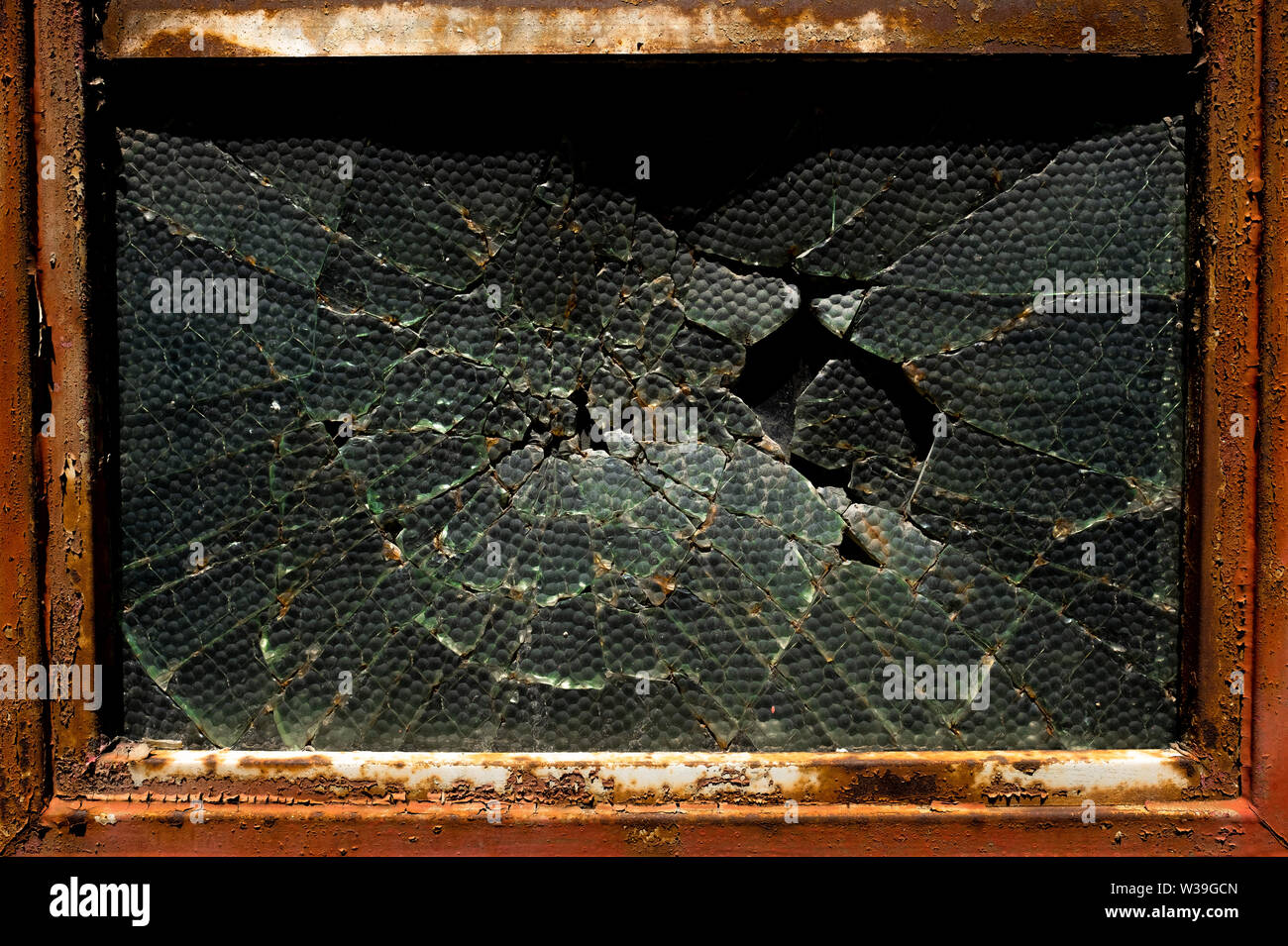Gros plan d'une fenêtre en verre texturé cassé dans un cadre en métal rouillé montrant des fissures radiales et la pourriture Banque D'Images