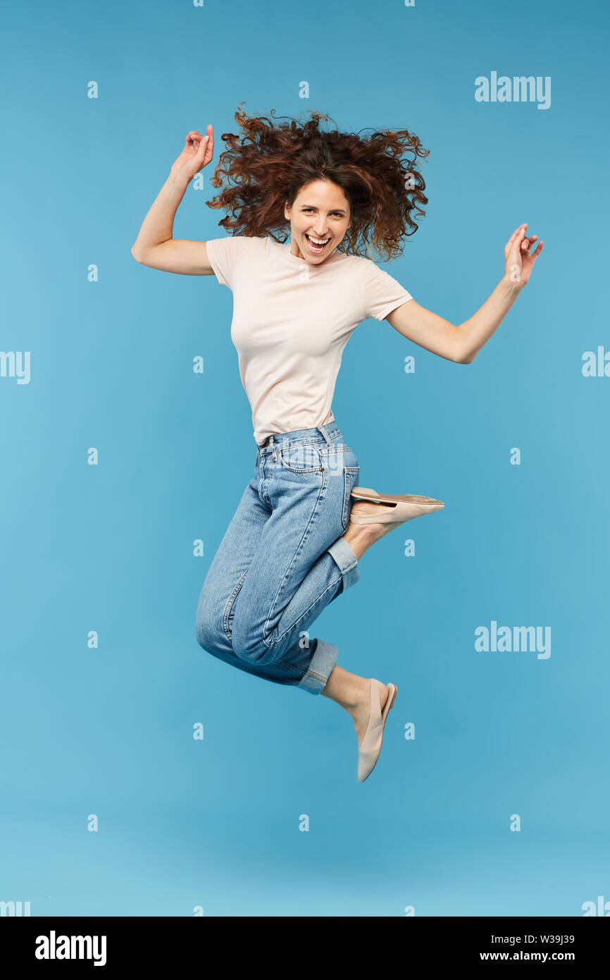 Jeune femme brune joyeuse en t-shirt blanc et jean bleu exprimant excitation tout en sautant dans l'isolement Banque D'Images