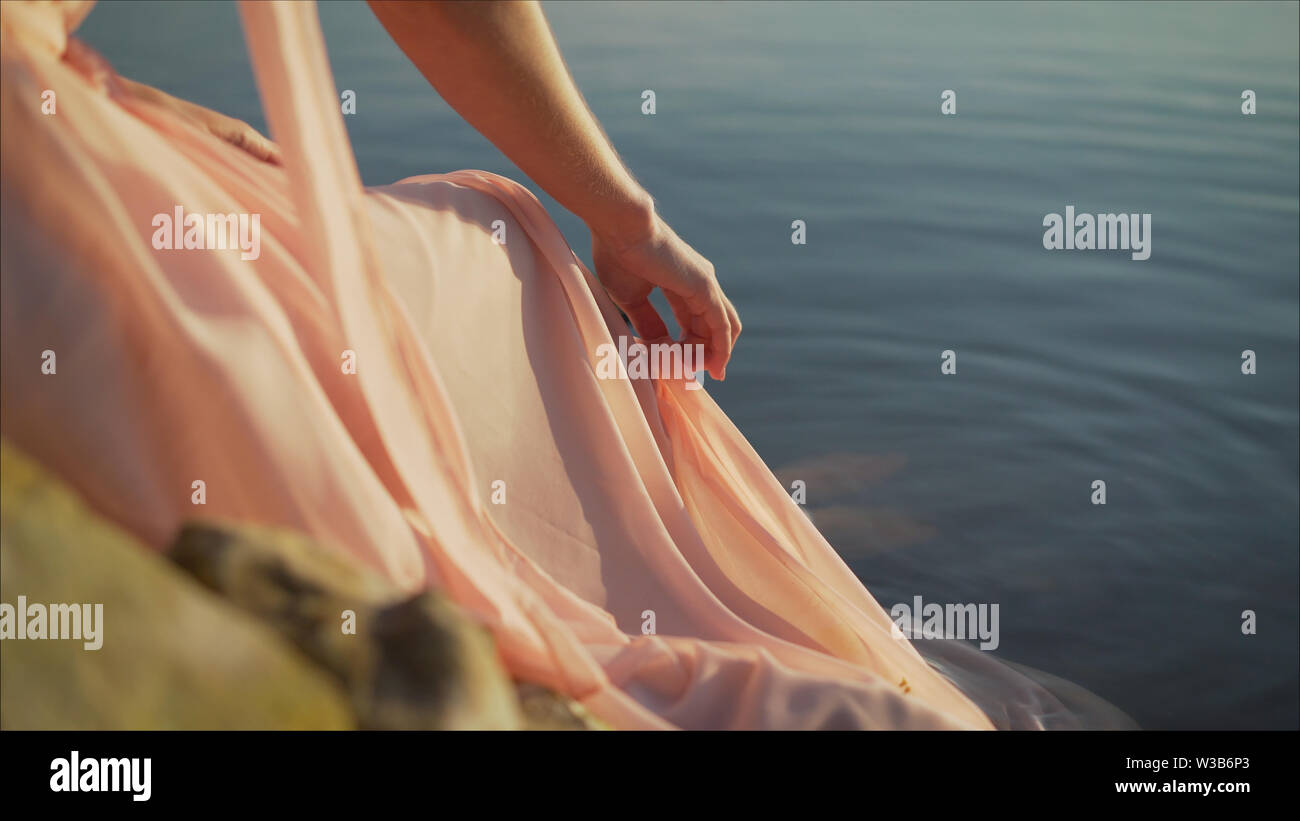Fille en robe rose pâle aux cheveux rouges et son sac à dos à marcher le long de la rivière, assis sur le sable. La jeune fille dans une robe rose sur la rive du fleuve. Banque D'Images
