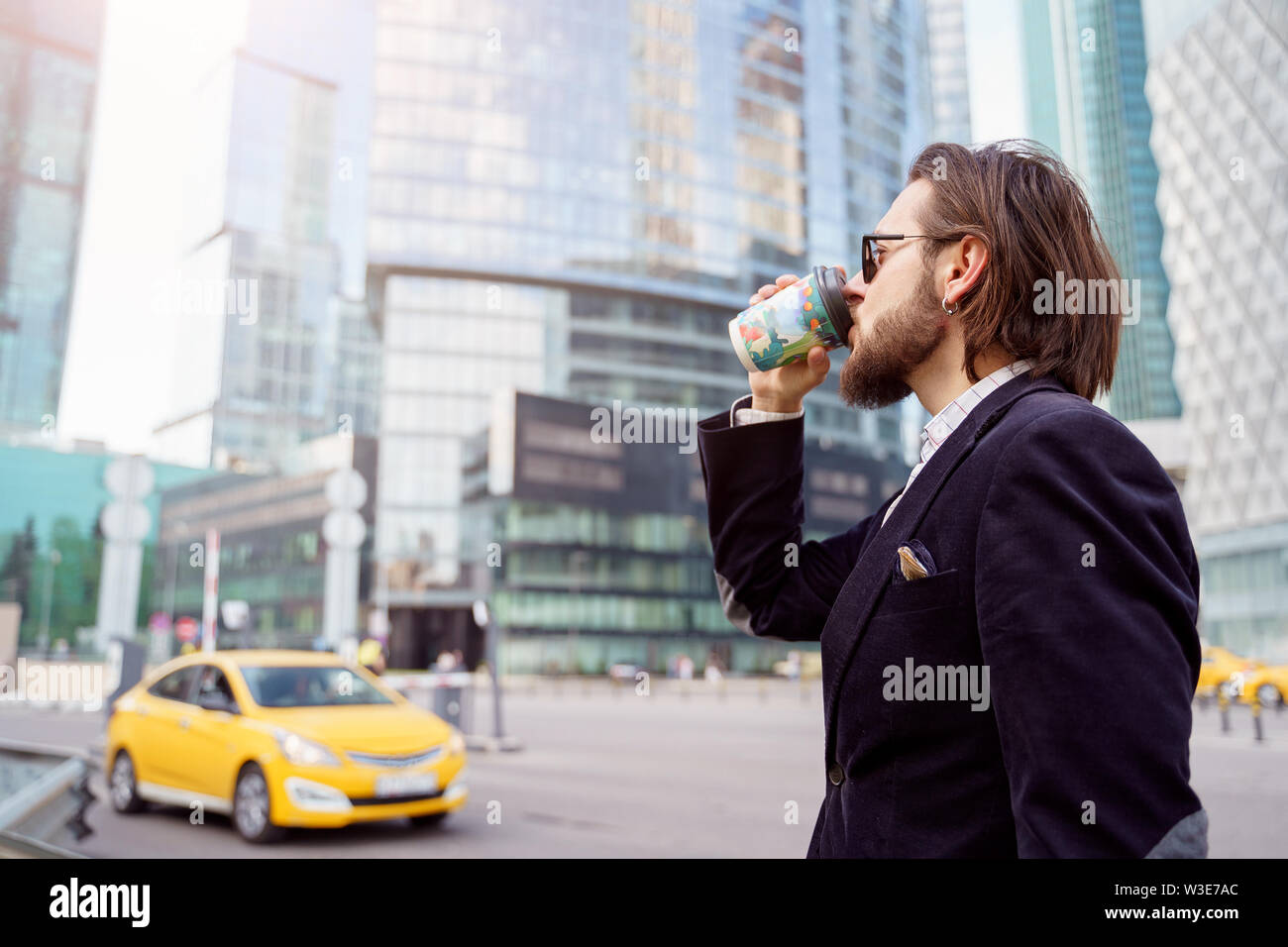 Photo sur côté de brunet avec barbe et d'oreille dans l'oreille de boire de verre en ville sur fond d'immeubles de grande hauteur, voiture jaune Banque D'Images