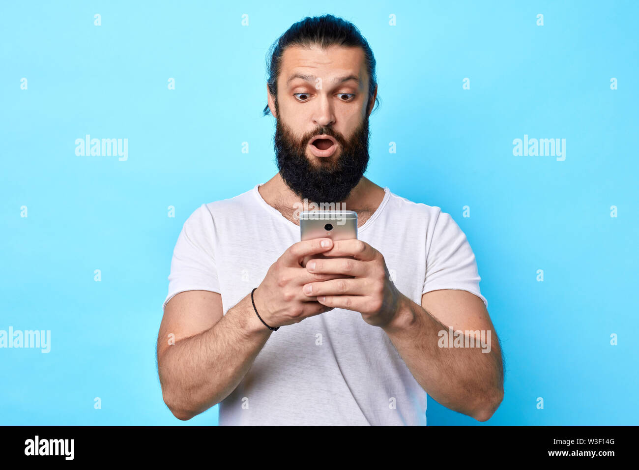 Jeune homme musclé fort choqué looking at mobile phone isolé sur fond bleu. l'homme est d'être choqué de recevoir des sms. close up portrait. studi Banque D'Images
