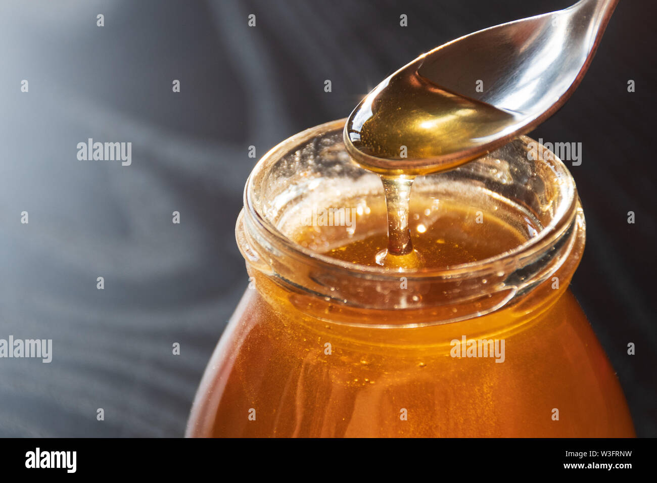 Délicieux miel bio naturels biologiques se déverse de la cuillère dans un pot de verre sur table en bois avec la réflexion du soleil, fond noir, Close up Banque D'Images