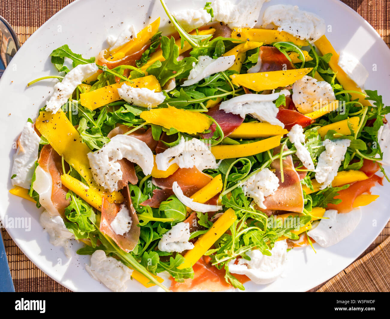 L'alimentation d'été : salade repas servi en plein air avec les tranches de mangue, roquette, mozzarella et jambon de parme sur une plaque blanche Banque D'Images