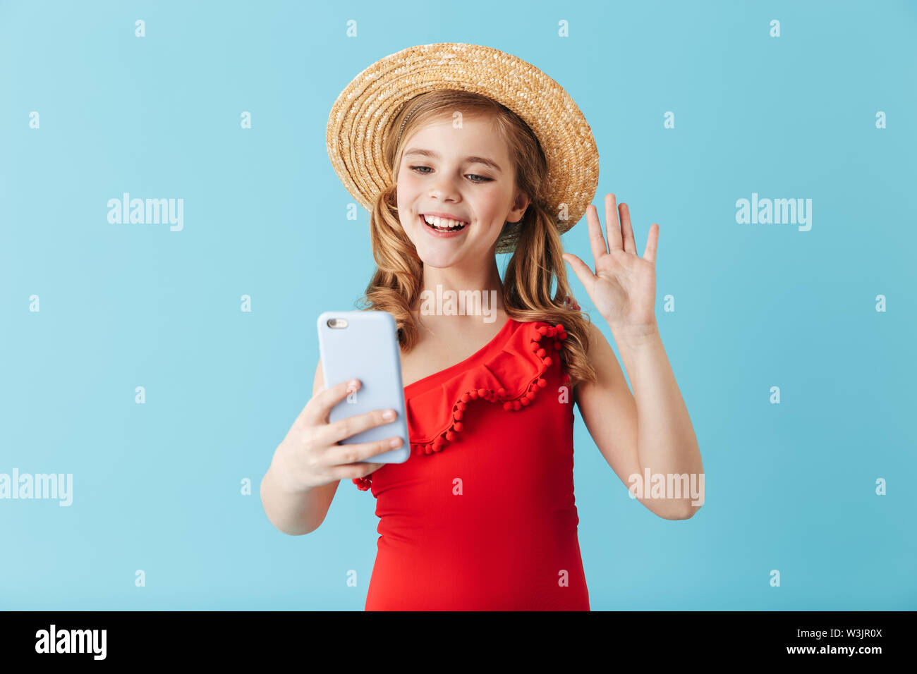 Happy little girl wearing hat standing d'été maillot de bain et isolé sur fond bleu, à l'aide de mobile phone Banque D'Images