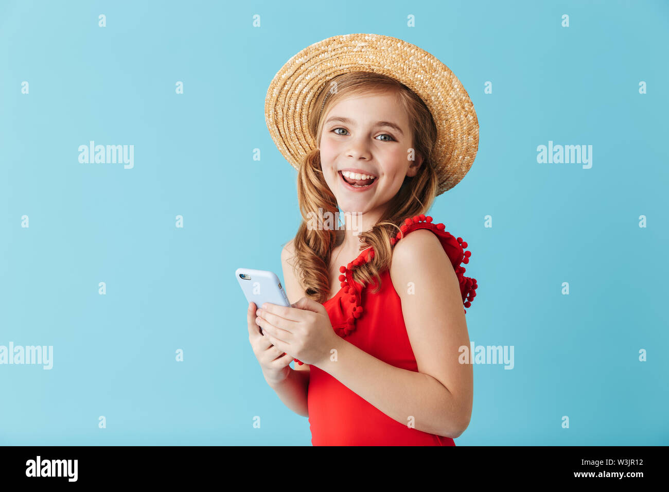 Happy little girl wearing hat standing d'été maillot de bain et isolé sur fond bleu, à l'aide de mobile phone Banque D'Images