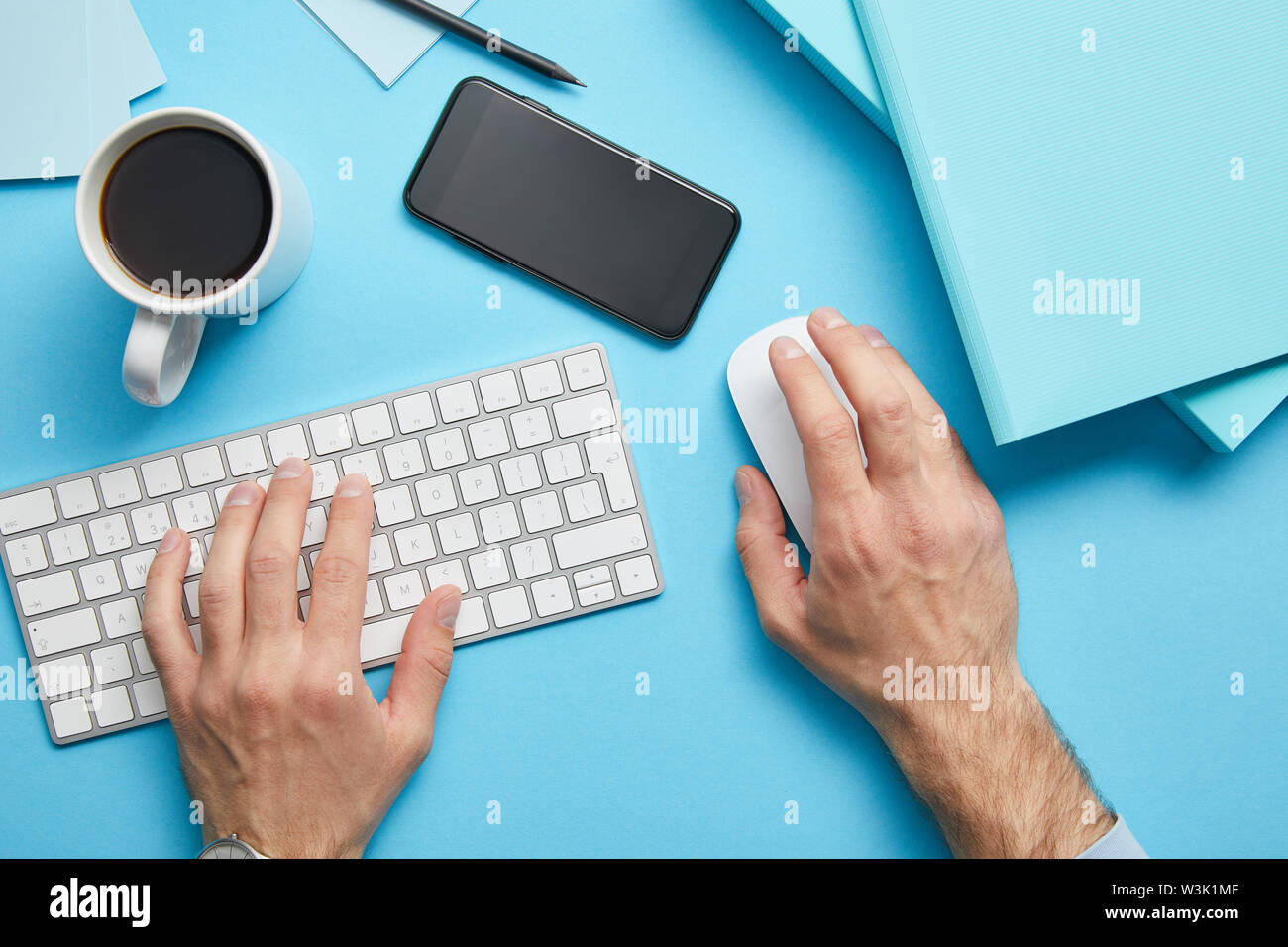 Portrait de l'homme à l'aide du clavier et souris de l'ordinateur sur le lieu de travail avec papiers, smartphone et tasse de café sur fond bleu Banque D'Images