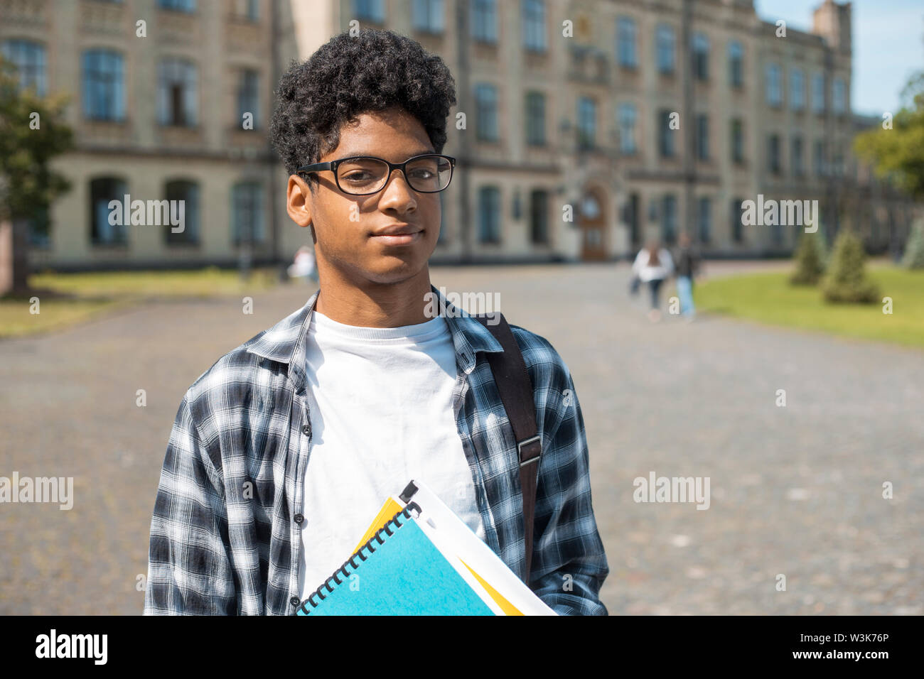 Un étudiant africain souriant avec des lunettes et des livres près de l'université. Portrait d'un jeune homme noir heureux debout sur un fond universitaire. Banque D'Images